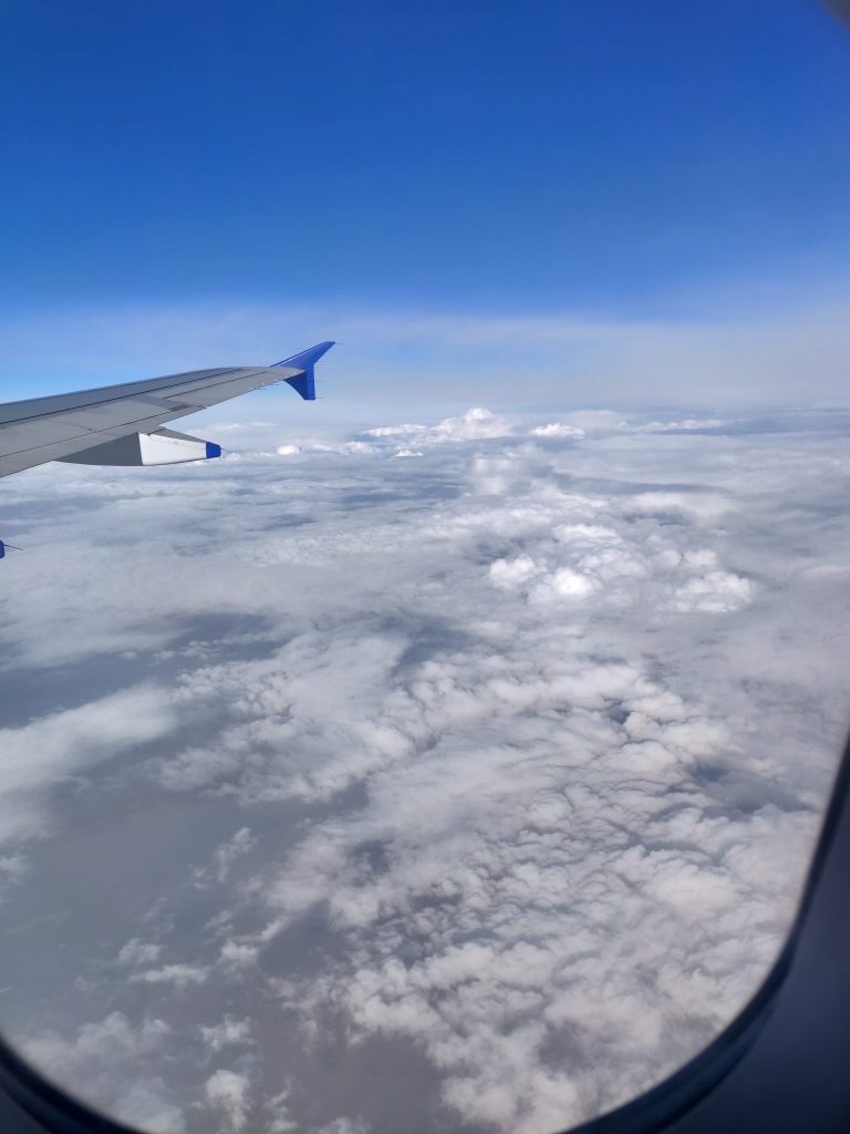 View from an airplane window showing a wing and a vast sea of fluffy white clouds below against a backdrop of clear blue sky.